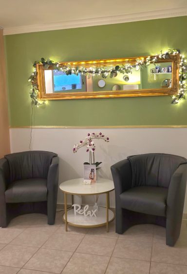 Wartebereich - 1- Zwei graue Sessel flankieren einen runden Tisch mit einer Orchideenblume vor einem grünen Wand.