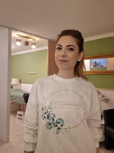 Kosmetikerin Frau mit blonden Haaren in einem weißen Sweatshirt mit Blumenmuster, lächelt im Raum.